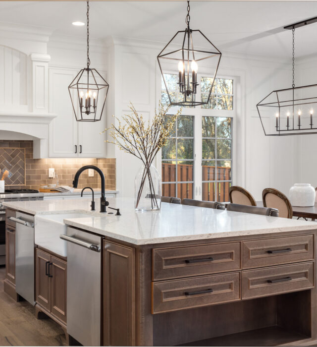 Modern kitchen with a large island featuring a white countertop and dark wood cabinets. Three geometric pendant lights hang above, and dining area is visible in the background.