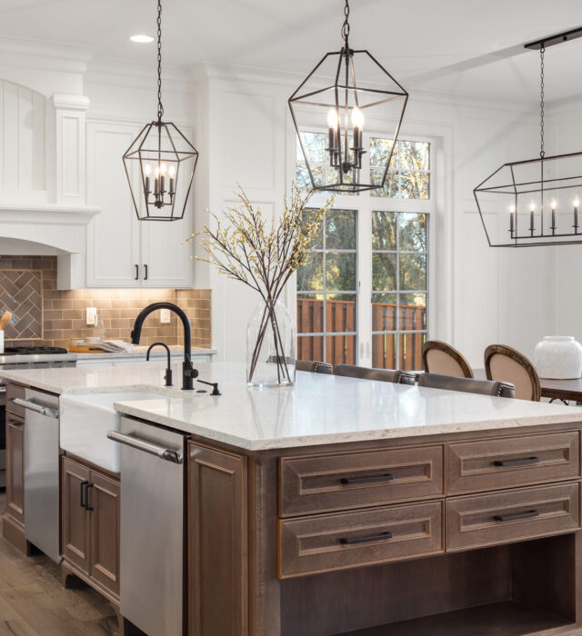 Modern kitchen with a large island featuring a white countertop and dark wood cabinets. Three geometric pendant lights hang above, and dining area is visible in the background.