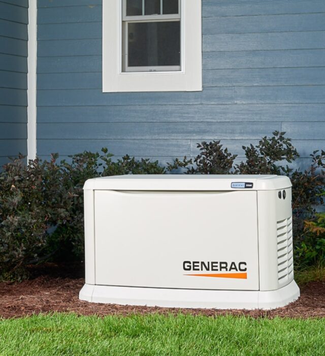 A whole-home Generac generator installed in by the bushes besides a blue house.