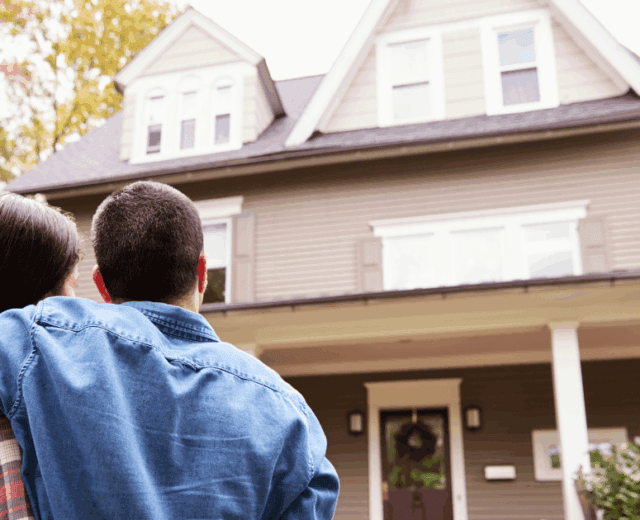 Two people stand with arms around each other, facing a large two-story house with a porch, seen from behind on a clear day.