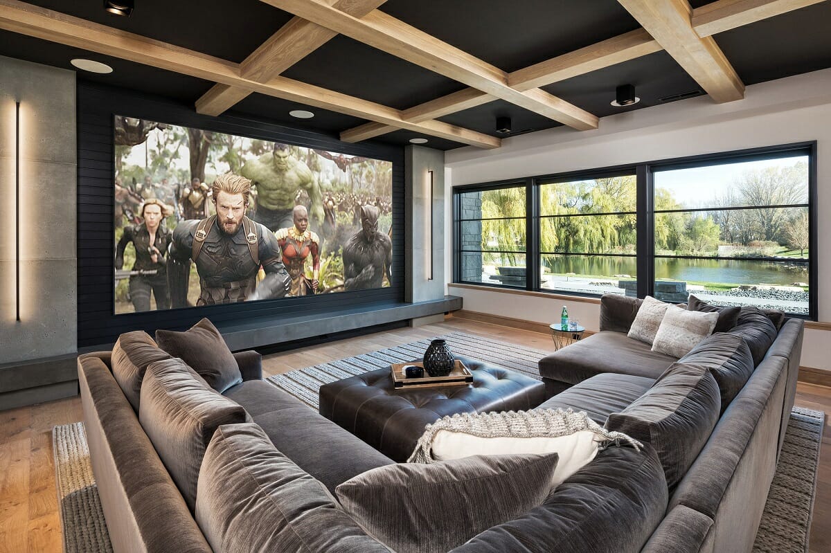 A modern home theater room features a large sectional sofa, a wall-mounted screen displaying a movie scene, and large windows overlooking a pond and trees.