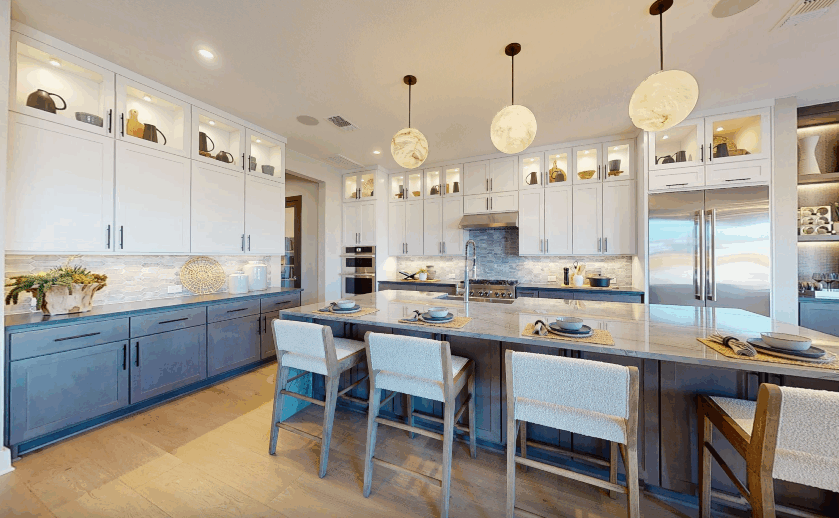 Modern kitchen with white upper cabinets, blue lower cabinets, a large island with four chairs, pendant lights, and stainless steel appliances.