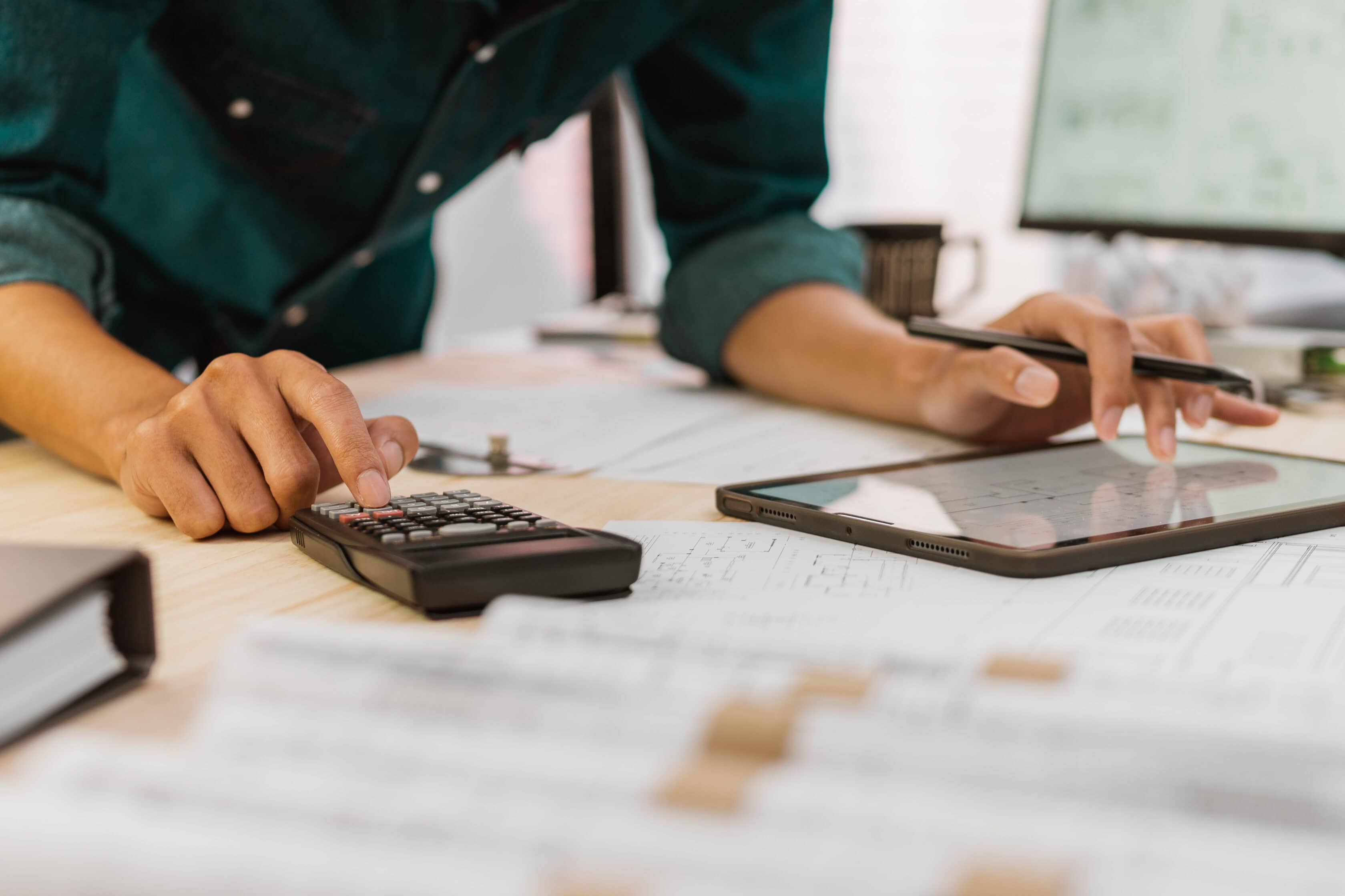 Person using a calculator and writing on a tablet at a desk with documents, suggesting financial or administrative work.