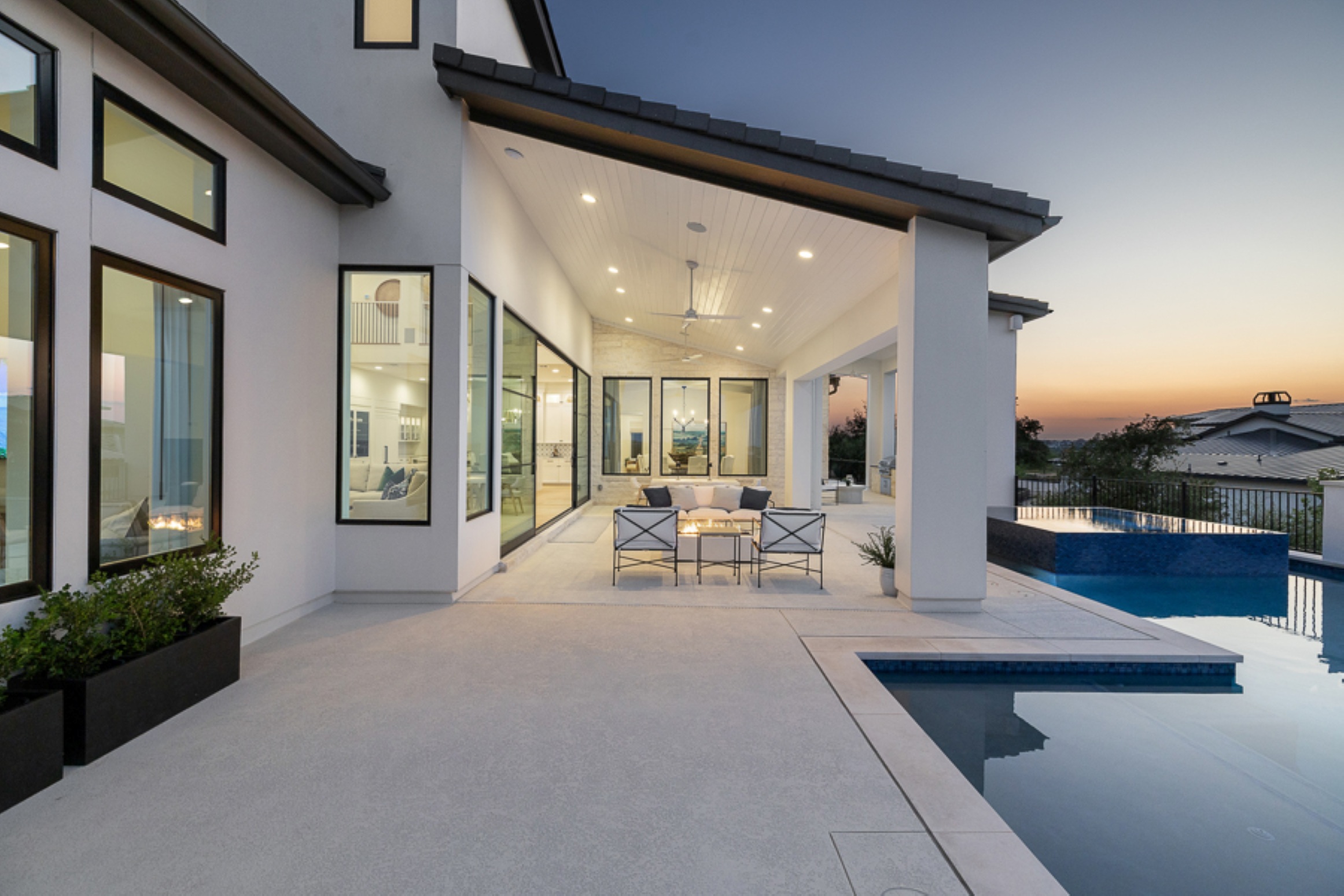 Modern house with large windows and outdoor patio, featuring a pool and seating area at sunset.