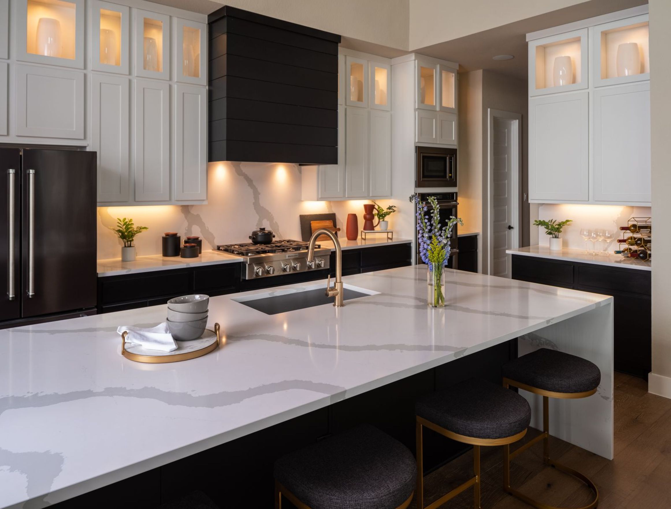 Modern kitchen with a large white marble island, black lower cabinets, white upper cabinets, built-in appliances, and decorative plants and vases on the counters and shelves.
