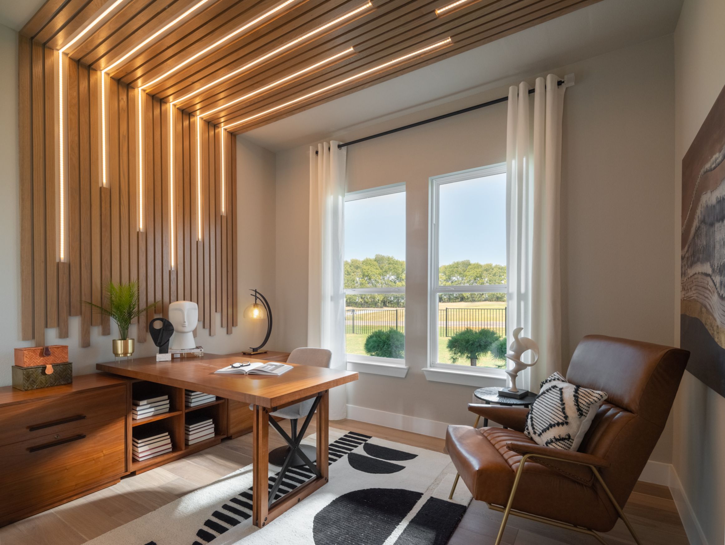 Modern home office with a wooden desk, leather chair, decorative wall lights, large windows with white curtains, and a patterned rug.