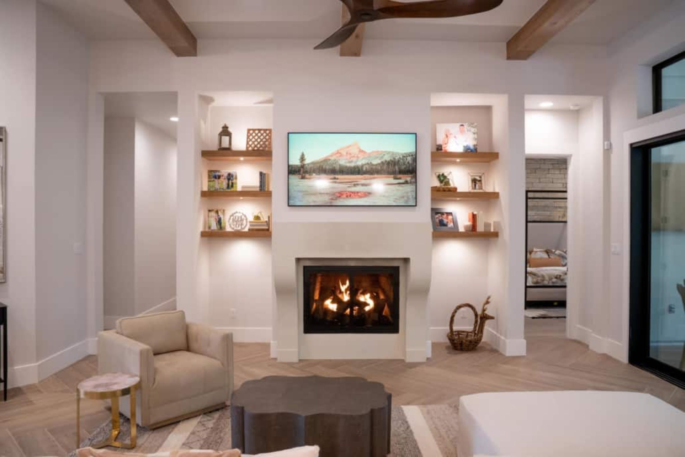 A modern living room with a lit fireplace, wall-mounted TV, built-in shelves with decor, and neutral-colored furniture on a wood-patterned floor.