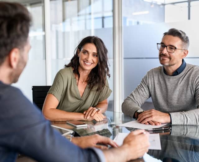 Three people sit at a glass table in an office, two facing one, with papers and smartphones on the table, engaged in a conversation.