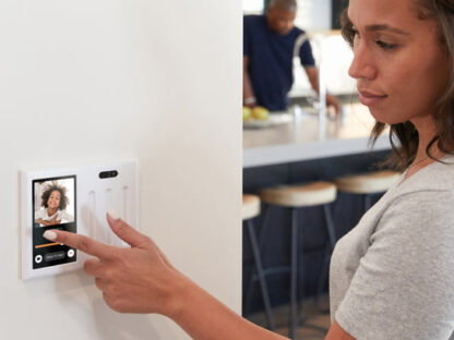 A woman uses a touchscreen wall device with a video call interface, displaying another woman’s face, in a modern home kitchen setting.