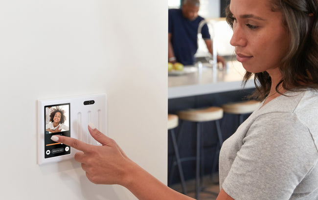 A woman uses a touchscreen wall device with a video call interface, displaying another woman’s face, in a modern home kitchen setting.