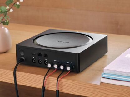 A black Sonos audio amplifier with various cables connected, sitting on a wooden table next to a vase with flowers and a stack of books.