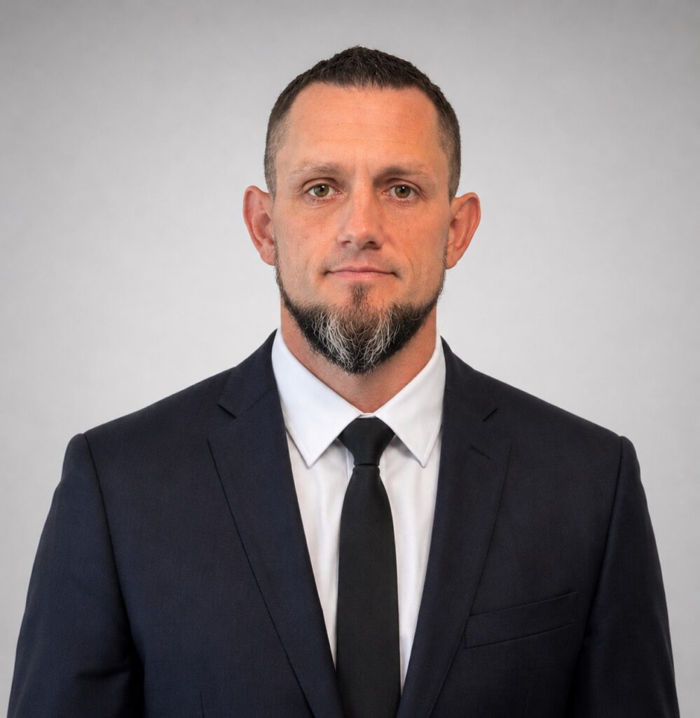 A man with short hair, a beard, and a mustache is wearing a dark suit, white shirt, and black tie, standing against a plain light gray background.
