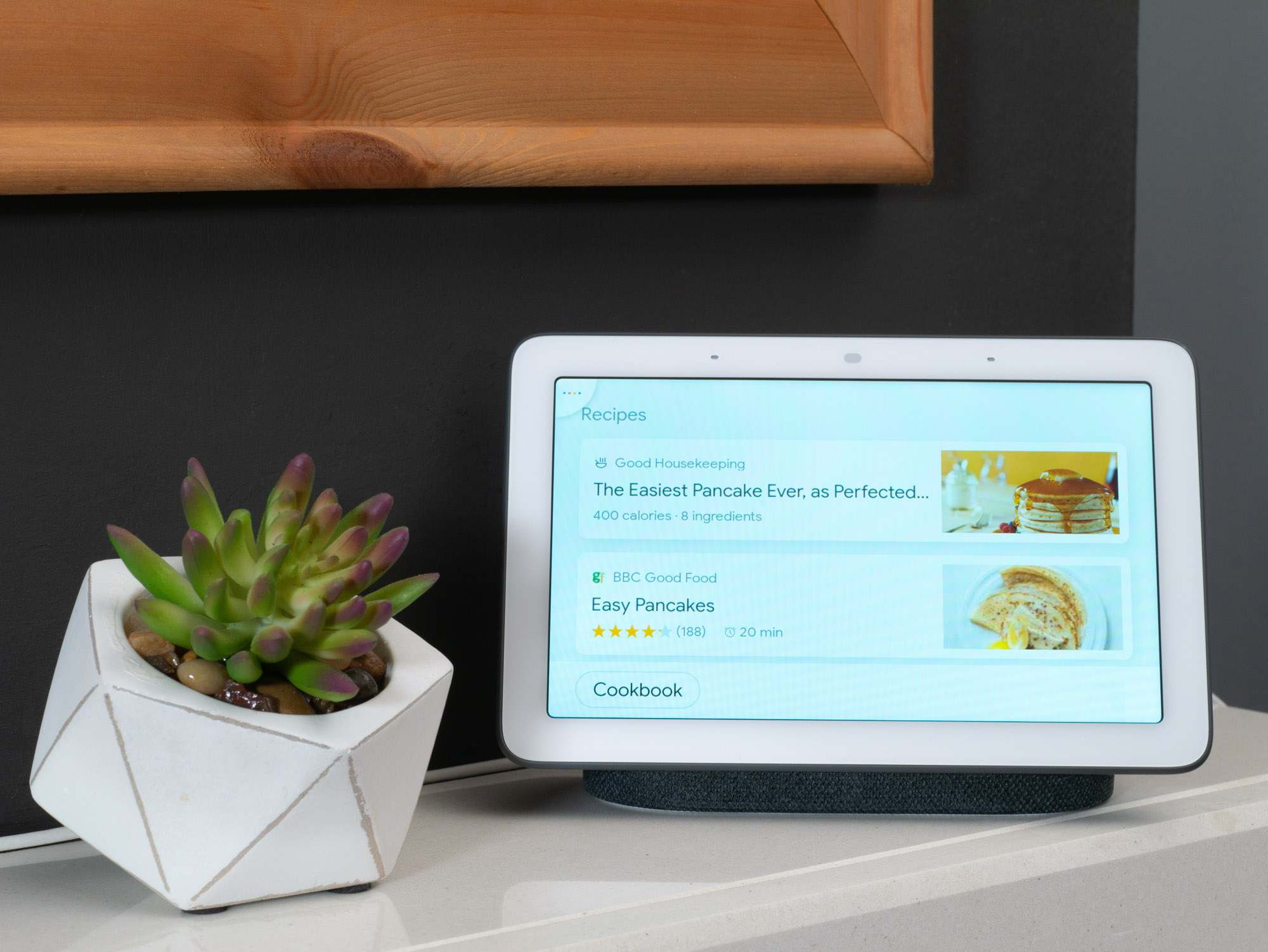 A smart display on a countertop shows pancake recipes next to a small potted succulent.