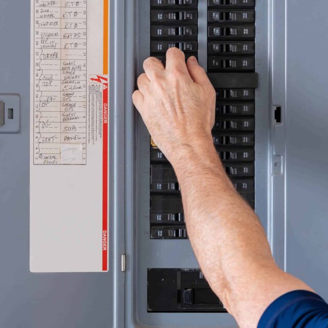 A person adjusts a breaker switch in an open electrical panel.|Electrical panel with wires in a wooden framework of a house under construction.|