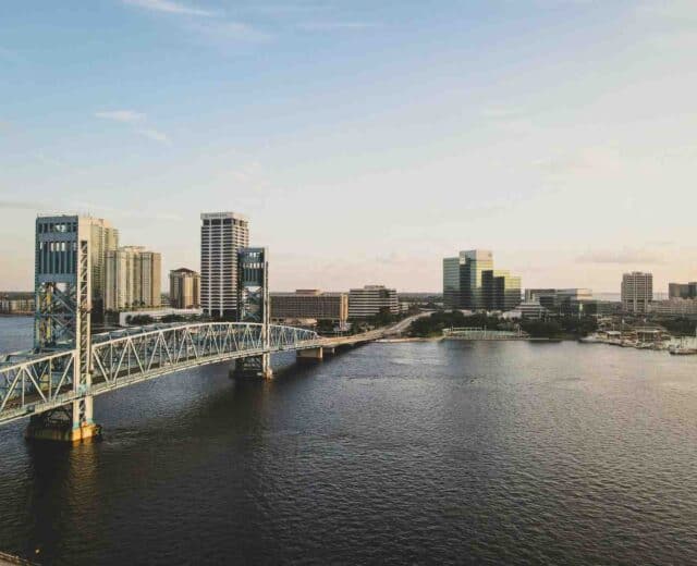 An aerial view of the Jacksonville, Florida skyline with the Main Street Bridge spanning the St. Johns River under a clear sky.